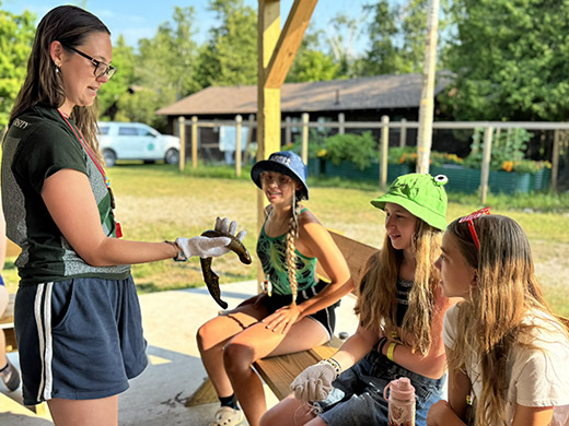 A photo of a teenage girl holding a live sea lamprey while three additional teenage girls seated on a bench look at the creature with curious looks.